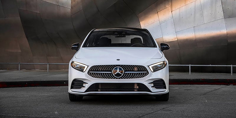 Front view of a white Mercedes-Benz A-Class sedan parked on asphalt in front of a metal architectural wall