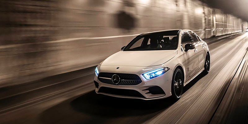 A white Mercedes-Benz A-Class sedan speeds leftward through a dark tunnel, headlights blazing