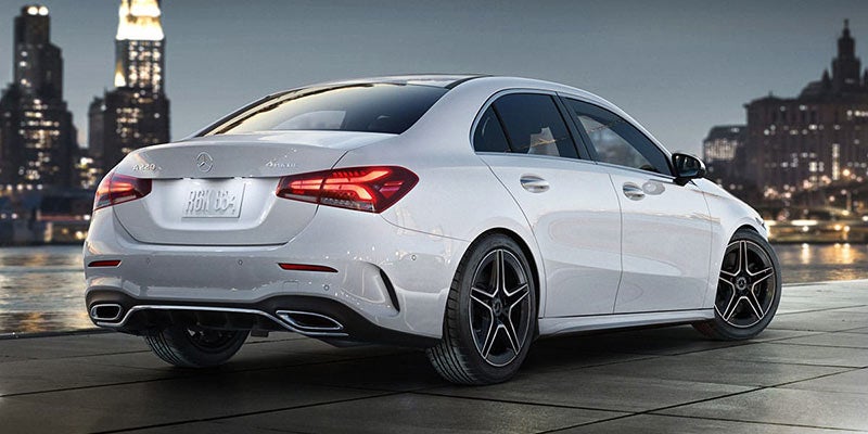 Rear view of a white Mercedes-Benz A-Class sedan with multi-spoke wheels, parked outdoors at dusk with city lights behind