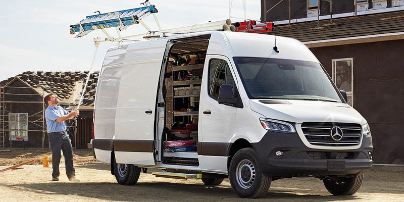 A man lowers a ladder from the roof rack of a white Mercedes-Benz Sprinter work van with its side door open.