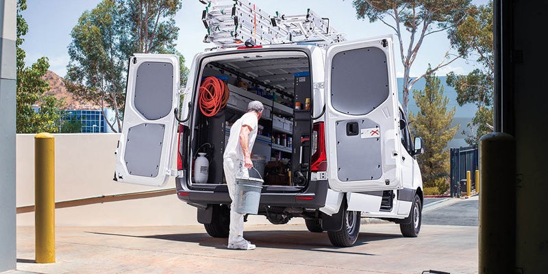 Man in white work clothes loads a bucket into the back of an open, ladder-equipped white work van.