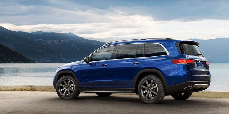Side view of a blue Mercedes-Benz GLS SUV parked on a paved road next to a lake surrounded by mountains
