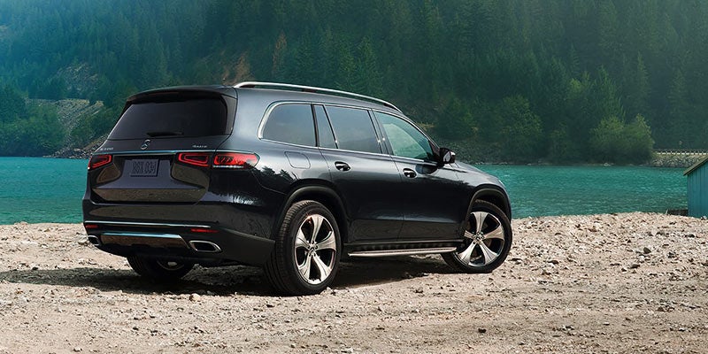 Dark grey Mercedes-Benz GLS SUV parked on a sandy shore by a river with green hills in the background