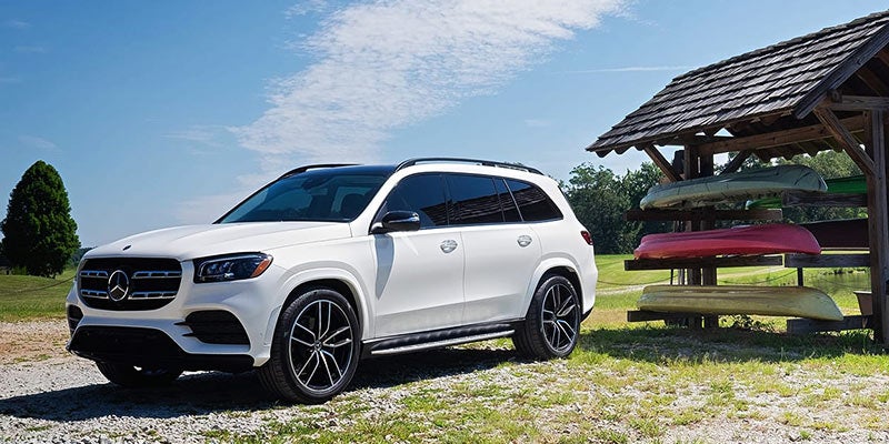 White Mercedes-Benz GLS SUV parked on gravel next to a wooden boathouse with stacked canoes and kayaks