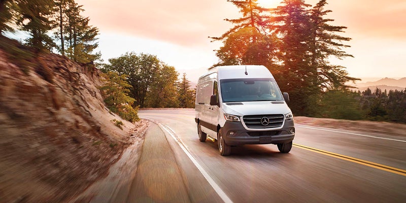 White Mercedes-Benz Sprinter van driving along a winding mountain road surrounded by trees during sunset