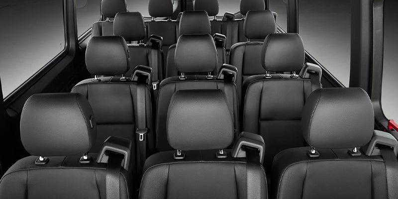 Interior view of a passenger van or bus, showing rows of dark gray or black seats with headrests and seatbelts.