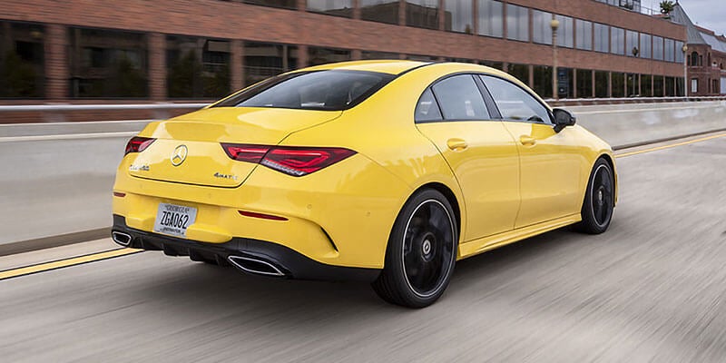 Rear three-quarter view of a yellow Mercedes-Benz sedan driving on a city street alongside office buildings