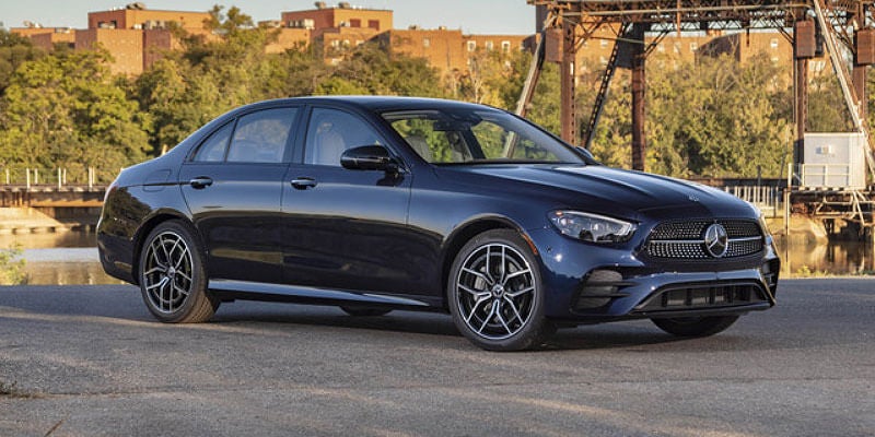 Dark blue Mercedes-Benz sedan parked on a paved waterfront area with trees and industrial structures in the background