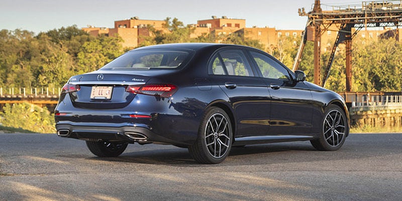 Rear three-quarter view of a dark blue Mercedes-Benz sedan parked beside a waterfront with industrial structures in the background