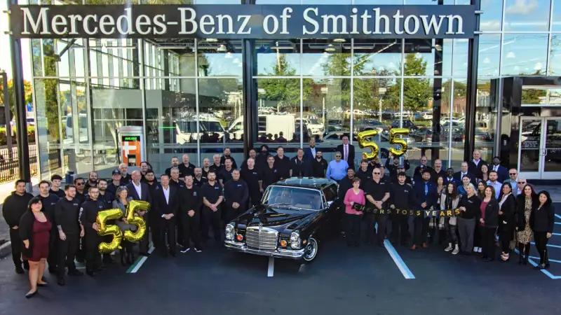 Group photo of over 50 staff in front of Mercedes-Benz of Smithtown, celebrating with a vintage black Mercedes and 55 balloons.