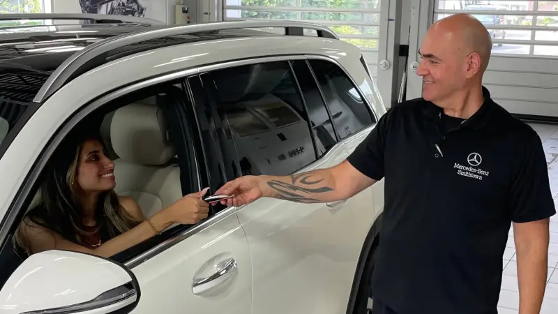 A customer in a white Mercedes-Benz receives keys from a smiling service employee.