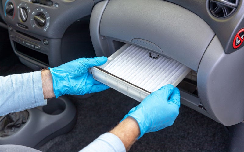 Hands in blue gloves removing a clean cabin air filter from a car's dashboard glove compartment