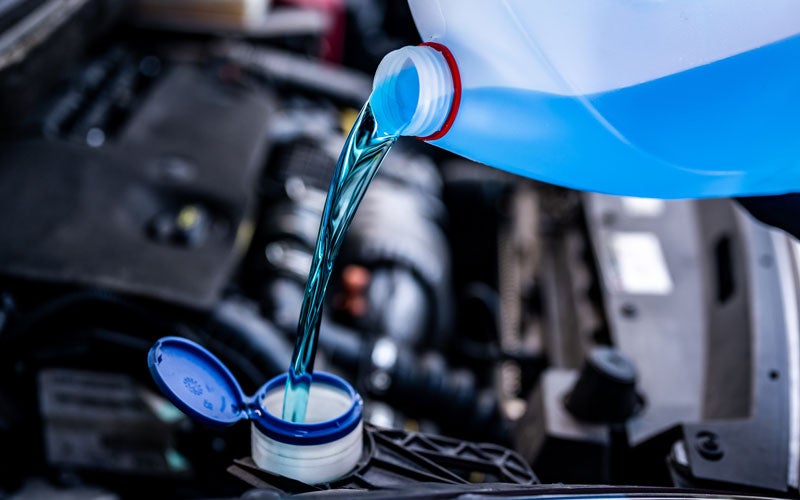 Blue windshield wiper fluid being poured from a transparent bottle into a car's reservoir under the hood
