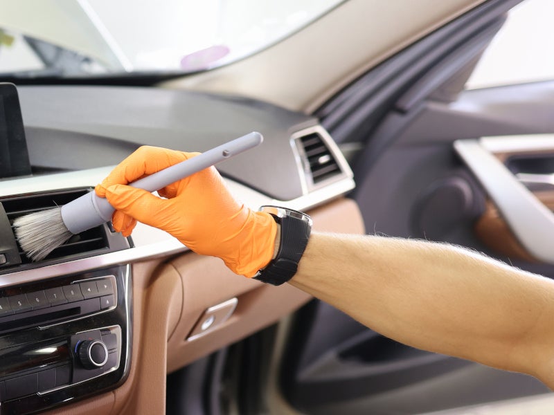 A person in orange gloves cleans a car's dashboard air vent with a detailing brush