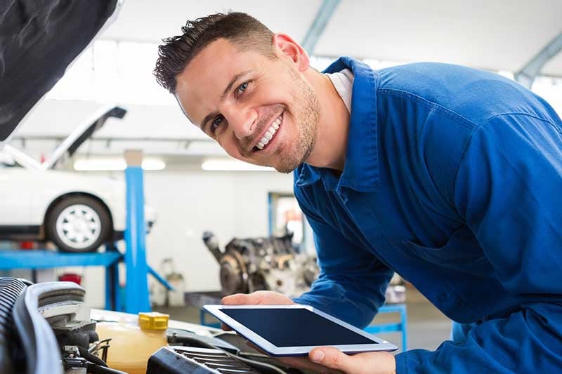 A smiling mechanic in a blue uniform leans over a car engine, holding a tablet while looking at the viewer